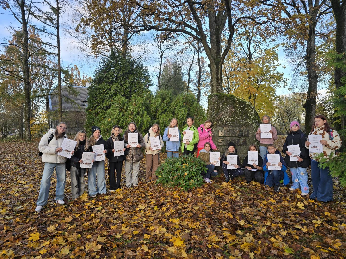 Jaunie mākslinieki ar pateicības rakstiem rokās pie Berģu skolas piemiņas akmens