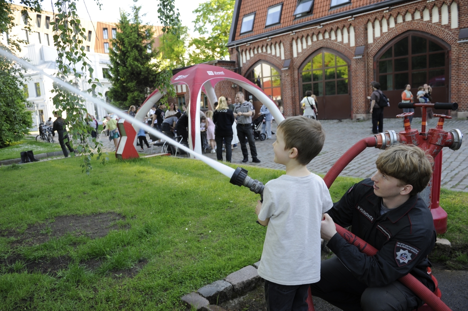VUGD nodarbinātais palīdz bērnam pieturēt ugunsdzēsības šļūteni. Fonā Latvijas Ugunsdzēsības muzejs, piepūšamā telts un cilvēki. 