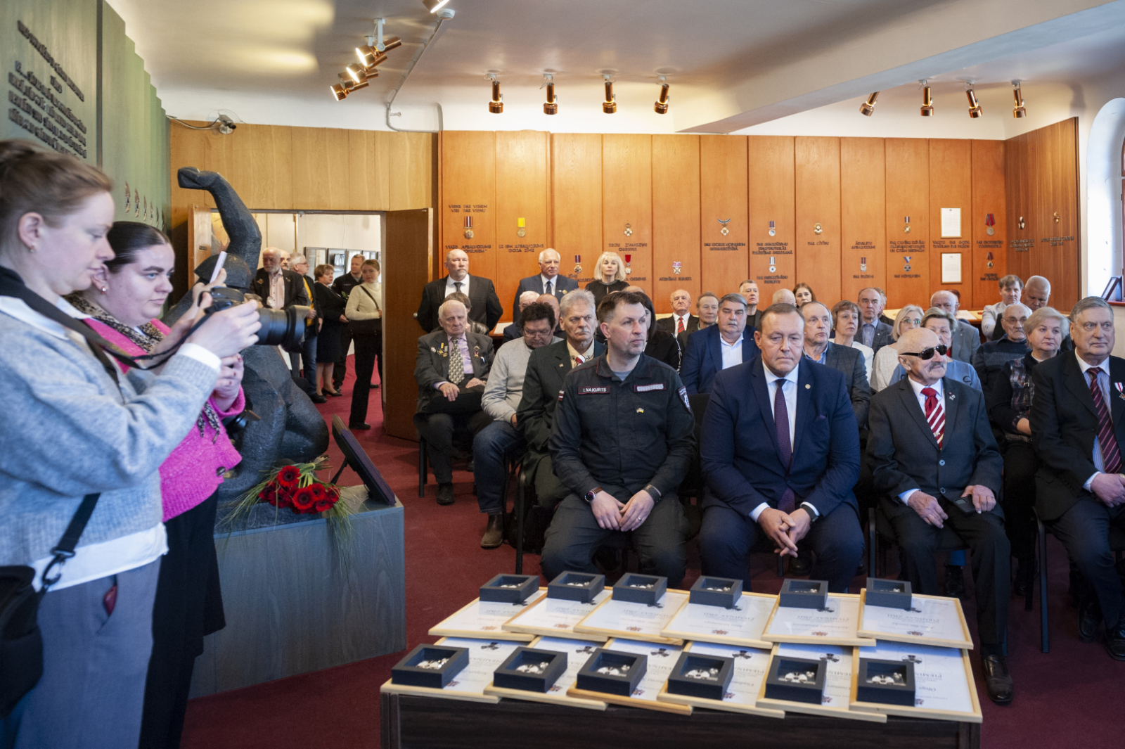 Latvijas ugunsdzēsības muzeja Slavas zālē sapulcējušies pasākuma viesi. Labajā pusē viesi sēž, kreisajā pusē stāv kājās.Priekšplānā uz galda izkāroti apbalvojumi. 
