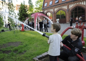 VUGD nodarbinātais palīdz bērnam pieturēt ugunsdzēsības šļūteni. Fonā Latvijas Ugunsdzēsības muzejs, piepūšamā telts un cilvēki. 