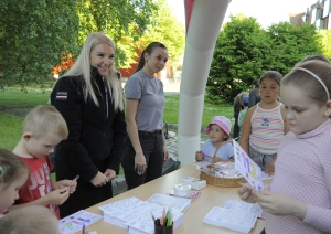 Divi VUGD nodarbinātie un bērni sastājušies ap galdu, uz kura izdales meteriāli - krāsojamās lapas ar uzdevumiem, zīmuļi.