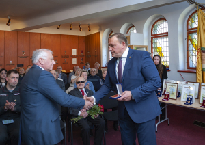 Iekšlietu ministrs Rihards Kozlovskis pasniedz piemiņas zīmi Černobiļas AES seku likvidēšanas dalībniekam. Fonā sanākušie pasākuma dalībnieki. Labajā pusē uz galda izkārtoti apbalvojumi.