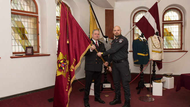 Slavas zālē divi vīrieši tur rokās karogu tumši sarkanā krāsā ar izšuvumiem, aizmugurē viņiem uzstādīti  divi  karogi un  virsnieku formas tērpi