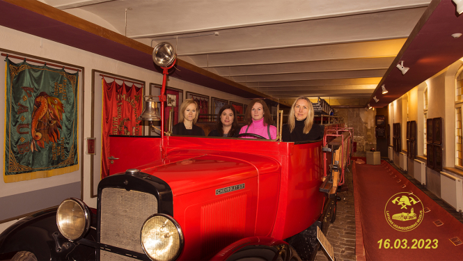 Chevrolet SIX ugunsdzēsēju mašīnas kabīnē sēž četras sievietes. Pa kreisi pie sienas brīvprātīgo ugunsdzēsēju biedrību karogi