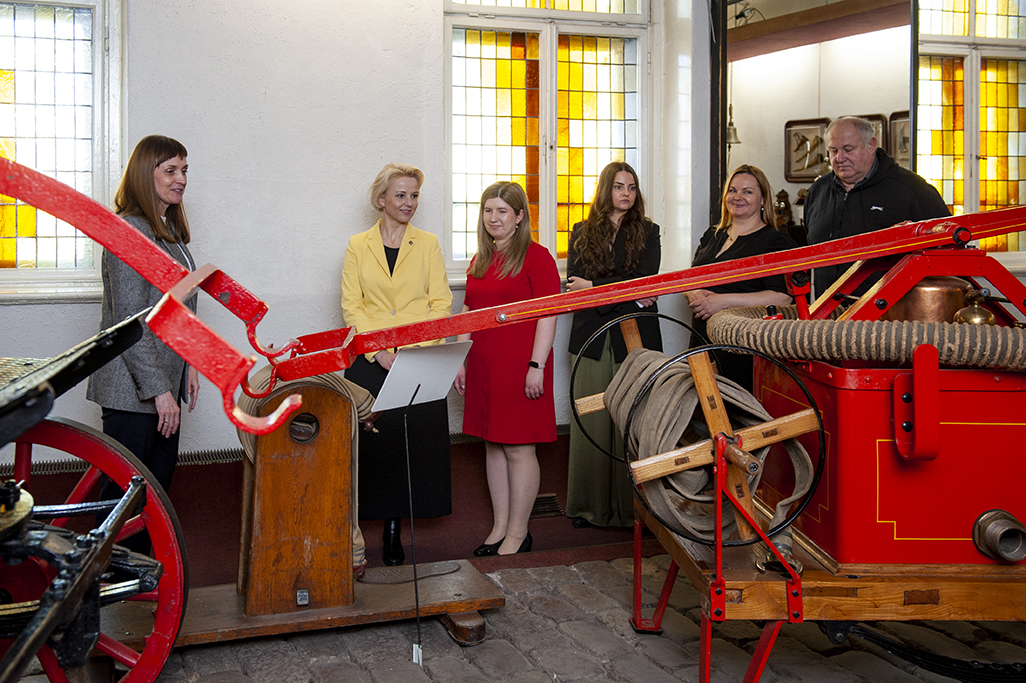 Priekšplānā Latvijas Ugunsdzēsības muzeja eksponāts, aiz tā seši cilvēki pēta to.