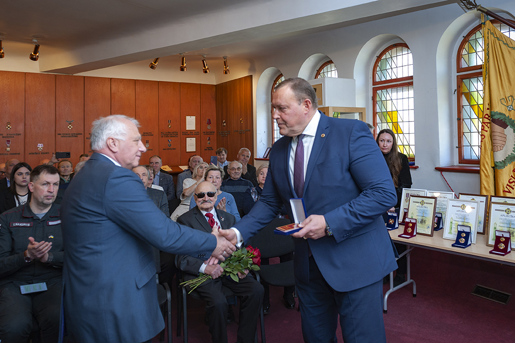 Iekšlietu ministrs Rihards Kozlovskis pasniedz piemiņas zīmi Černobiļas AES seku likvidēšanas dalībniekam. Fonā sanākušie pasākuma dalībnieki. Labajā pusē uz galda izkārtoti apbalvojumi.