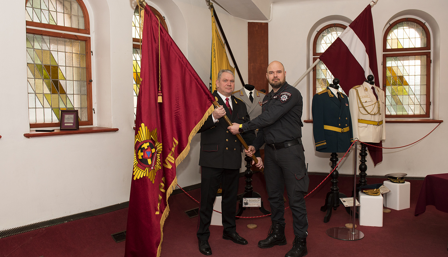 Slavas zālē divi vīrieši tur rokās karogu tumši sarkanā krāsā ar izšuvumiem, aizmugurē viņiem uzstādīti  divi  karogi un  virsnieku formas tērpi